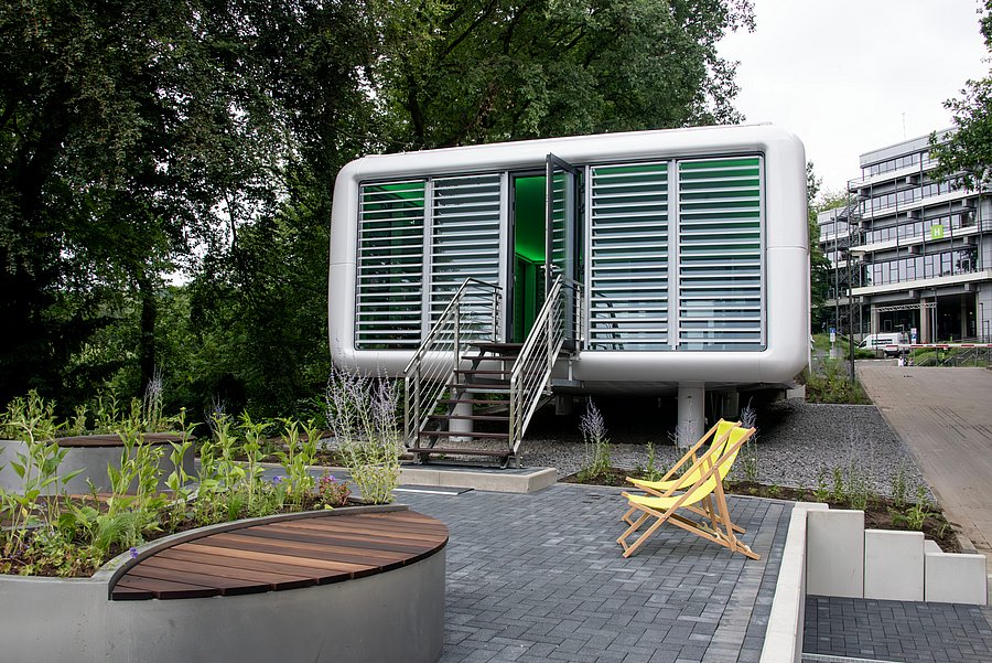 Das Foto zeigt einen kleinen, modernen, freistehenden Pavillon auf einem begrünten Außengelände. Das Gebäude ist rechteckig mit stark abgerundeten Ecken und einer hellgrau-weißen, glatten Außenhülle. Die gesamte Vorderseite besteht aus großen Fensterflächen, vor denen waagerechte Lamellen angebracht sind. In der Mitte befindet sich eine geöffnete Glastür. Durch sie fällt grünes Licht aus dem Innenraum nach außen. Zum Eingang führt eine kurze Metalltreppe mit etwa fünf Stufen und beidseitigen Edelstahlgeländern. Der Pavillon steht leicht erhöht auf Stützen; darunter und seitlich liegt grauer Kies. Im Vordergrund befindet sich ein gepflasterter Aufenthaltsbereich. Rechts vor dem Gebäude stehen zwei gelbe Liegestühle aus Holz und Stoff nebeneinander. Links und im unteren Bildbereich sind bepflanzte Hochbeete mit verschiedenen Gräsern, Stauden und violett blühenden Pflanzen zu sehen. In eines der Beete ist eine geschwungene Sitzfläche aus dunklen Holzlatten integriert. Große, dicht belaubte Bäume rahmen die linke und obere Bildhälfte ein und lassen die Umgebung sehr grün wirken. Rechts im Hintergrund steht ein mehrstöckiges, grau-weißes Universitätsgebäude mit langen Fensterreihen, Balkonen und einer außenliegenden Metalltreppe. Ein gepflasterter Weg führt daran vorbei. Insgesamt wirkt der Ort ruhig, modern und von viel Grün umgeben.