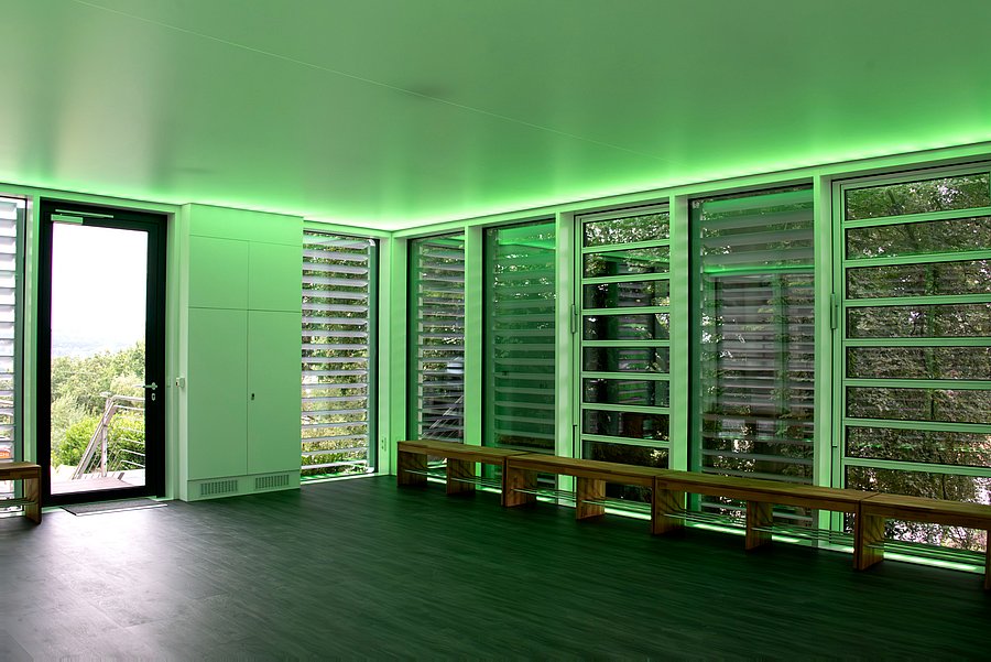 Innenansicht eines modernen, nahezu leeren Raumes mit großen Fensterflächen auf zwei Seiten. Der Raum hat einen dunklen, grau-schwarzen Boden, weiße Wände und eine helle, glatte Decke. Entlang der Deckenkanten verläuft eine indirekte grüne Beleuchtung, die den gesamten Raum in ein deutliches grünes Licht taucht. Die rechte Wand besteht fast vollständig aus bodentiefen Fenstern mit hellen Metallrahmen. Vor beziehungsweise hinter den Fenstern befinden sich waagerechte Lamellen, durch die dichtes grünes Laub und Baumkronen im Außenbereich zu sehen sind. Entlang dieser Fensterfront stehen mehrere schlichte, lange Holzbänke ohne Rückenlehne. Eine weitere kleine Holzbank ist teilweise am linken Bildrand zu erkennen. Links befindet sich eine dunkle Glastür, durch die Tageslicht in den Raum fällt. Hinter der Tür sind eine Außentreppe beziehungsweise ein Geländer sowie Bäume und eine hügelige Landschaft in der Ferne sichtbar. Rechts neben der Tür befindet sich ein großer, geschlossener weißer Einbauschrank. Zwischen Schrank und Fensterfront setzen sich weitere schmale Fenster mit horizontalen Lamellen fort.